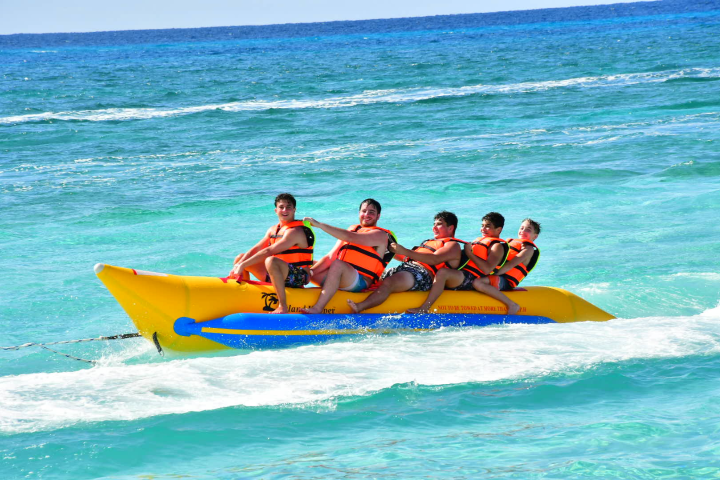 Five people wearing life vests ride a banana boat on the ocean.