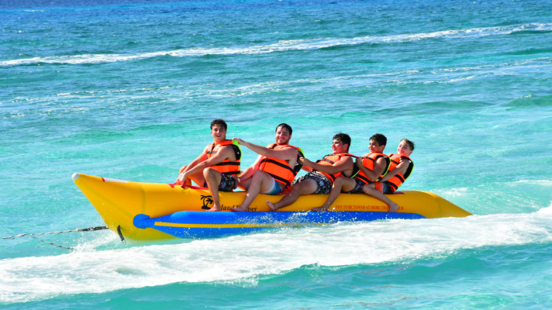 Five people wearing life vests ride a banana boat on the ocean.
