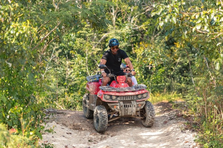 Man riding a red ATV on a forest trail surrounded by green trees.