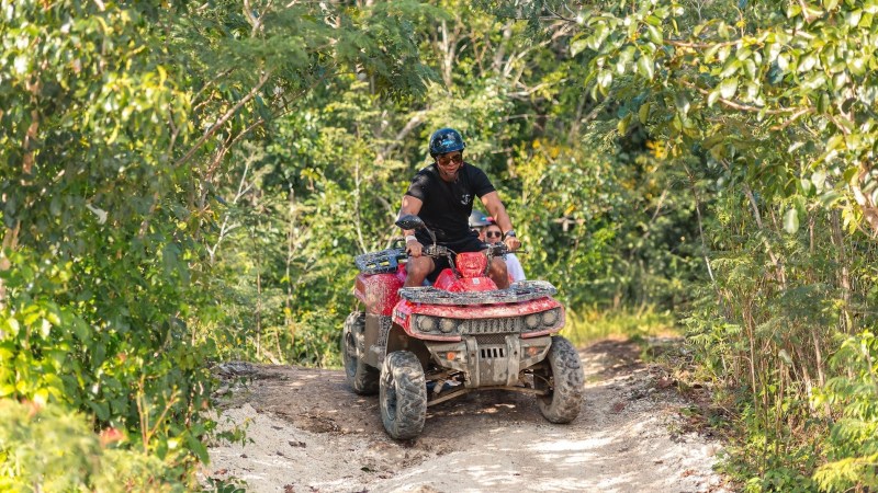 Man riding a red ATV on a forest trail surrounded by green trees.