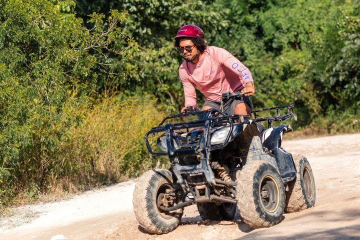Person in pink shirt and helmet riding ATV on dirt path with trees in background.