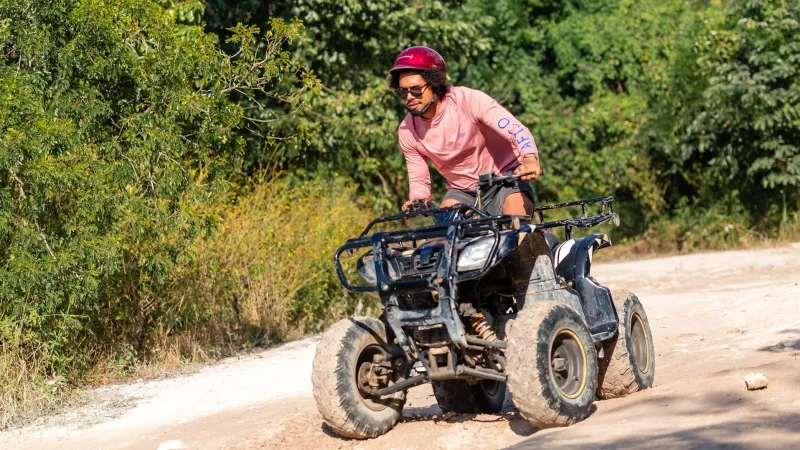Person in pink shirt and helmet riding ATV on dirt path with trees in background.
