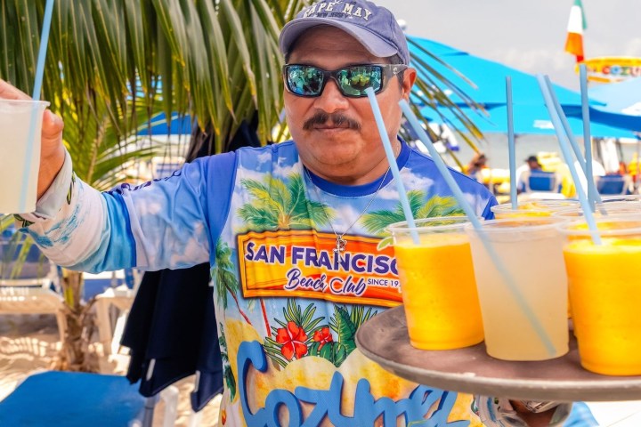 A man in a colorful shirt holds a tray of tropical drinks on a sunny beach under palm trees.