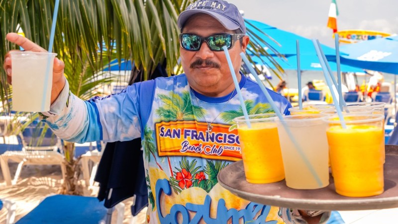 A man in a colorful shirt holds a tray of tropical drinks on a sunny beach under palm trees.