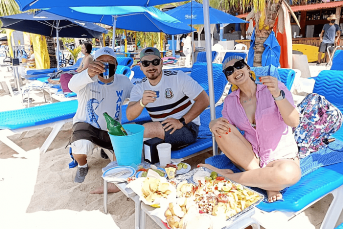Three people on beach chairs toast drinks, with food on a table under blue umbrellas.