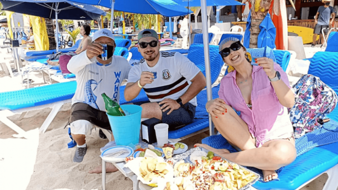 Three people on beach chairs toast drinks, with food on a table under blue umbrellas.
