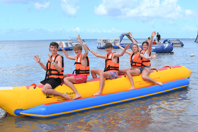 Five people on an inflatable banana boat, wearing life jackets, with arms raised, near an aquatic playground.
