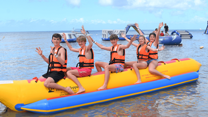 Five people on an inflatable banana boat, wearing life jackets, with arms raised, near an aquatic playground.