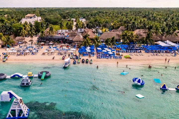 Aerial view of a beach resort with blue umbrellas, lounge chairs, and inflatable water park in the sea.