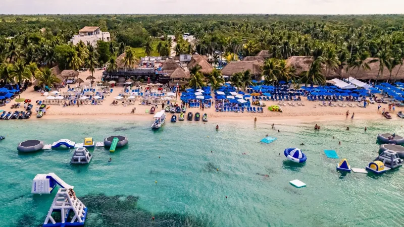 Aerial view of a beach resort with blue umbrellas, lounge chairs, and inflatable water park in the sea.