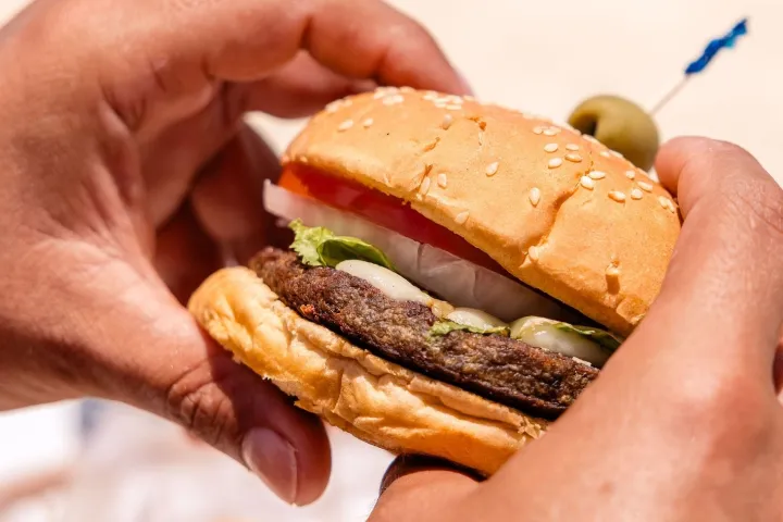 Close-up of hands holding a hamburger with lettuce, tomato, and onion on a sesame seed bun.