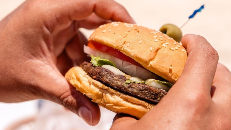 Close-up of hands holding a hamburger with lettuce, tomato, and onion on a sesame seed bun.