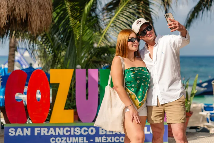 A couple takes a selfie in front of colorful 'Cozumel' sign at a beach.