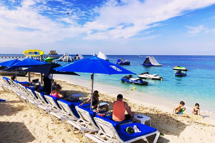 Beach scene with blue umbrellas, lounge chairs, jet skis, and a floating play structure in the sea.