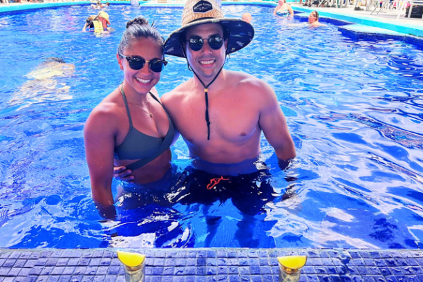 Smiling couple in sunglasses and hats standing in a pool near two drinks on the edge.