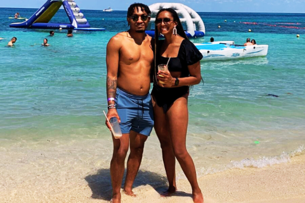 Couple stands on a beach, smiling with drinks in hand, inflatable water toys in background.
