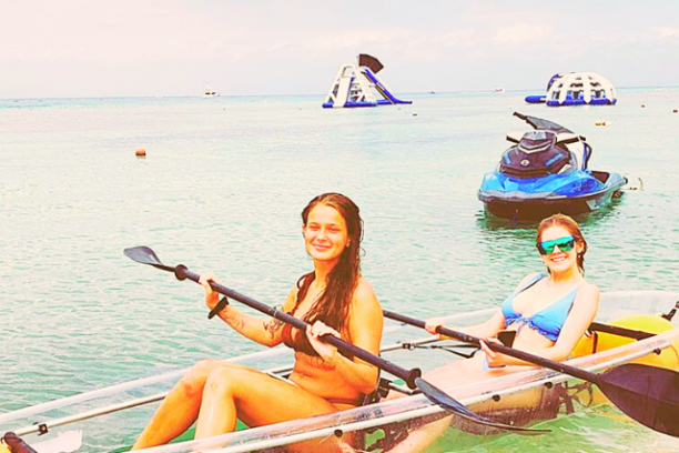 Two women kayaking in clear water with inflatable slides and jet ski in the background.