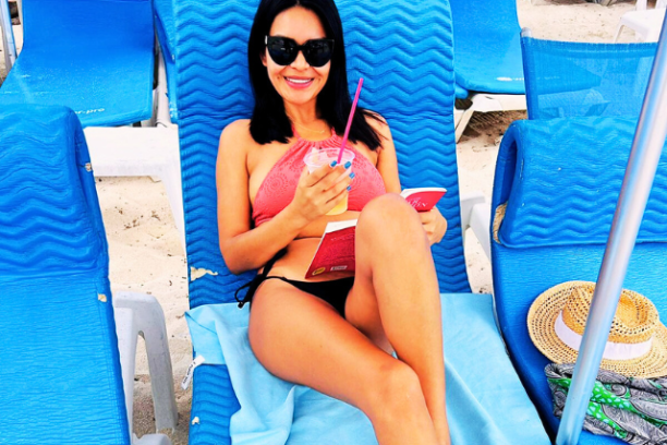Woman on beach lounge chair, holding a drink and a book, wearing sunglasses and a pink bikini.