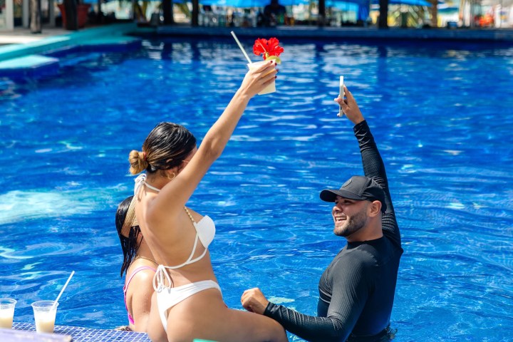 People in a pool raising drinks, smiling and taking photos, with a bright blue water background.