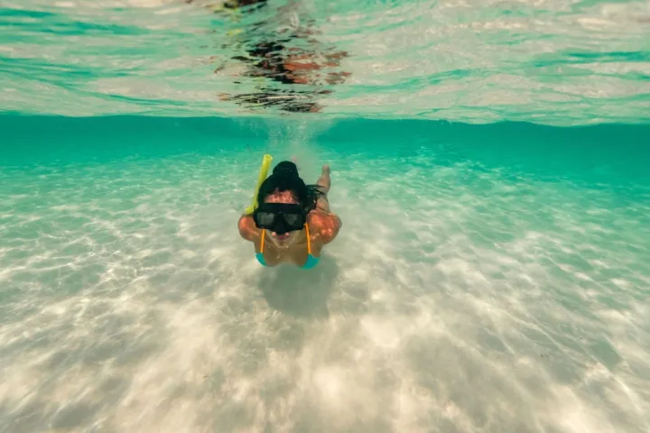 Person snorkeling underwater in clear, shallow ocean water with sandy bottom.
