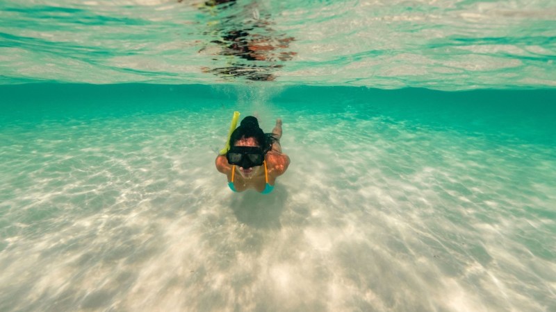 Person snorkeling underwater in clear, shallow ocean water with sandy bottom.
