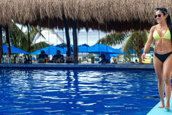 Woman in bikini walking by a pool with a thatched-roof bar in the background.