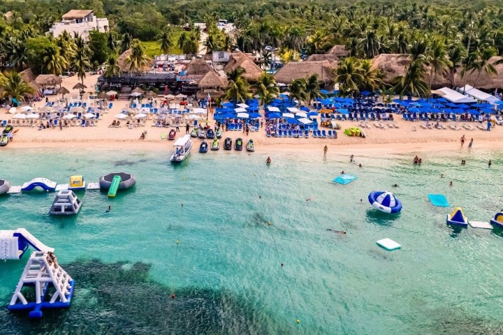 Beach with umbrellas, chairs, inflatables in water, and palm trees in background.