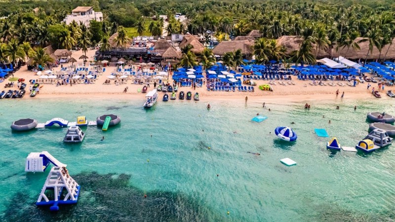Beach with umbrellas, chairs, inflatables in water, and palm trees in background.