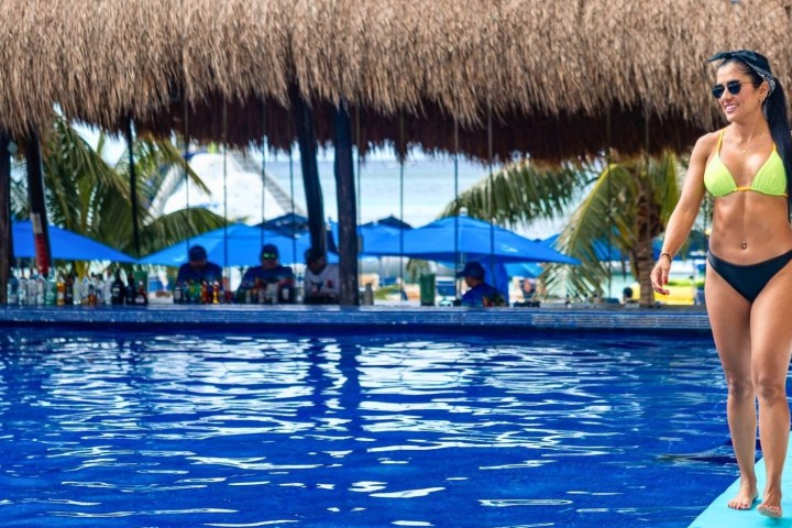 Woman in bikini walking by a poolside bar with straw roof and blue umbrellas.