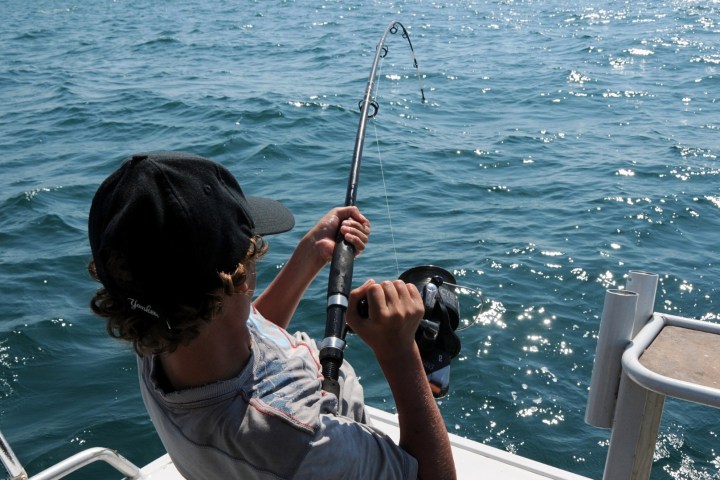 Person on a boat fishing, holding a fishing rod over the sparkling ocean.