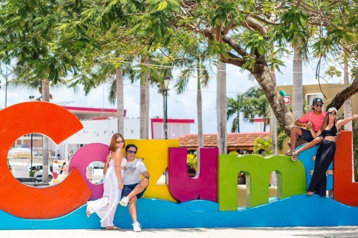Four people pose playfully on colorful 'Cozumel' sign with trees and buildings in the background.