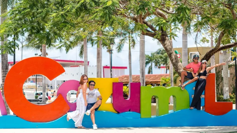 Four people pose playfully on colorful 'Cozumel' sign with trees and buildings in the background.