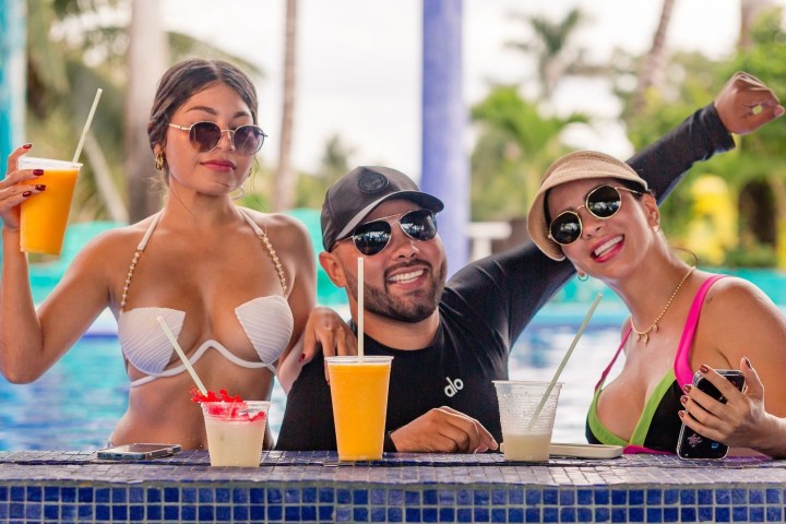 Three people at a poolside bar with drinks, wearing sunglasses and smiling.