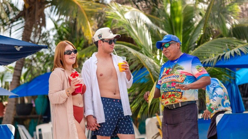 Three people on a beach holding drinks, surrounded by tropical trees and umbrellas.