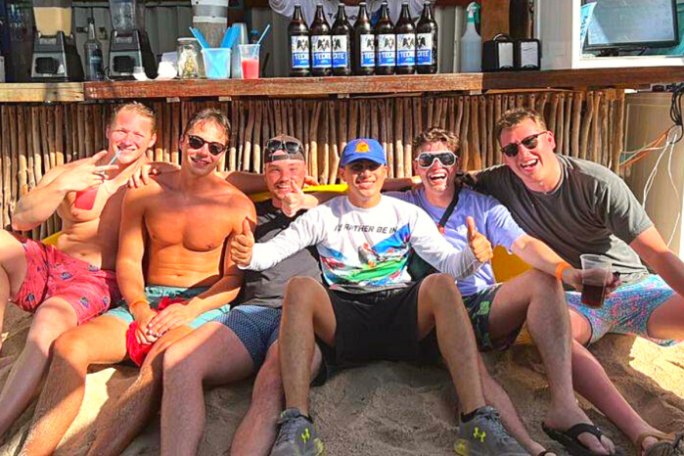 Six men sitting on sandy beach in front of a bar, smiling and giving thumbs up.