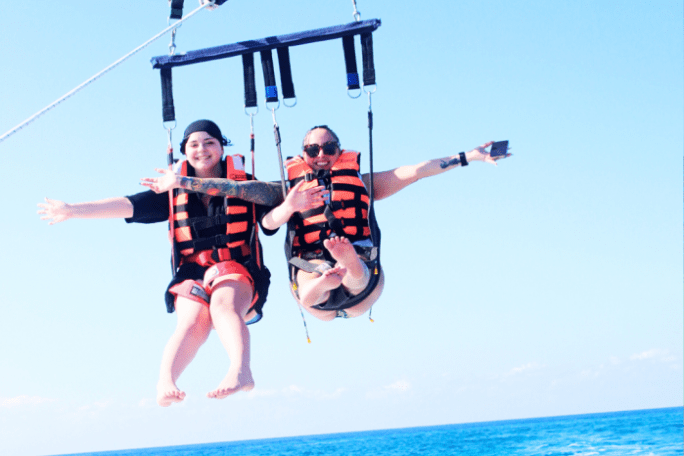 Two people parasailing over the ocean, wearing orange life jackets, smiling with arms outstretched.