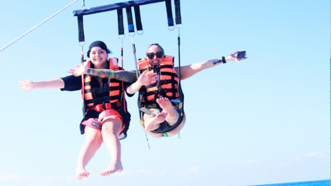 Two people parasailing over the ocean, wearing orange life jackets, smiling with arms outstretched.