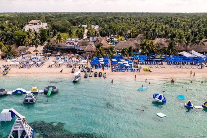Aerial view of a beach resort with blue umbrellas, palm trees, and water inflatables in the ocean.