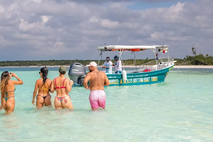 Four people walking in shallow ocean water towards a tour boat under a cloudy sky.