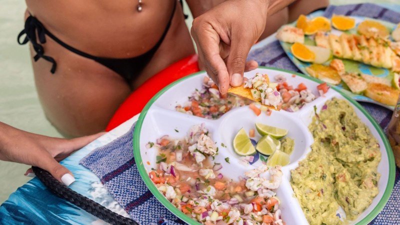 Person in a swimsuit holding a chip over a platter of dips, including guacamole and lime wedges, by the pool.