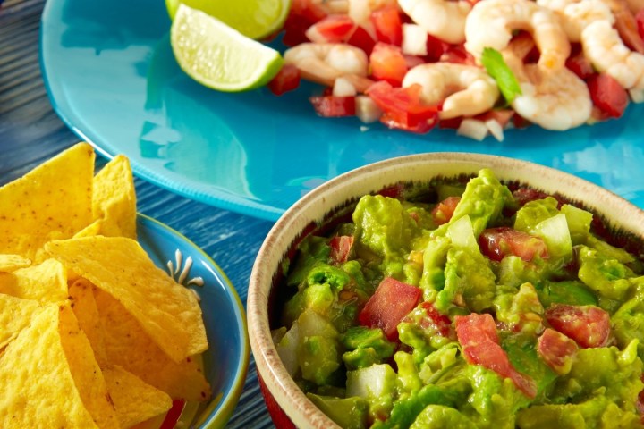 Guacamole in a bowl with nacho chips and a shrimp dish on a blue plate.