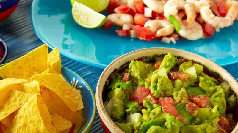 Guacamole in a bowl with nacho chips and a shrimp dish on a blue plate.