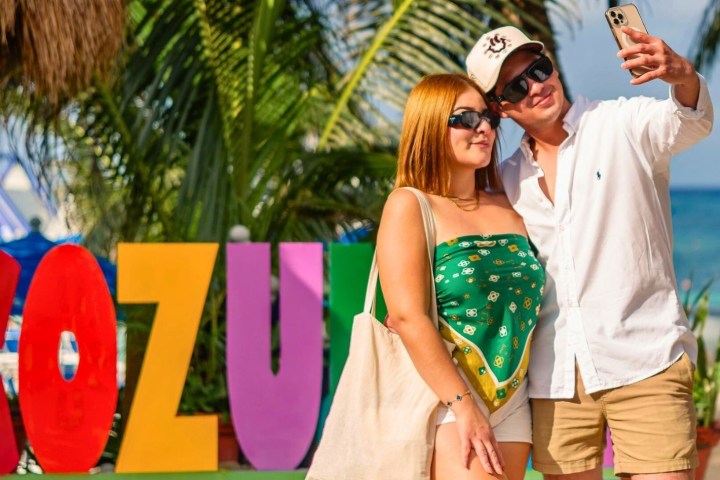 Two people taking a selfie in front of colorful 'Cozumel' letters on a tropical beach.