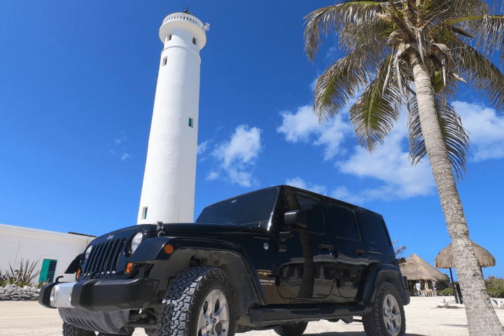 Black SUV parked near a tall white lighthouse and a palm tree on a sunny beach.