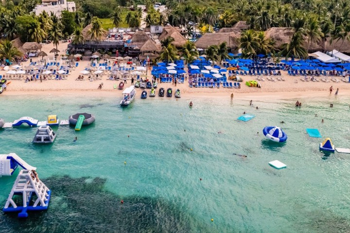 Beach with blue umbrellas, lounge chairs, and water park inflatables in the ocean.