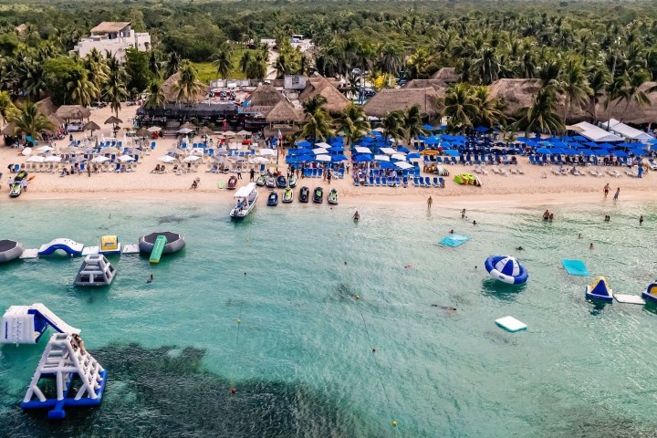 Beach scene with umbrellas, loungers, inflatable water park, and palm trees.