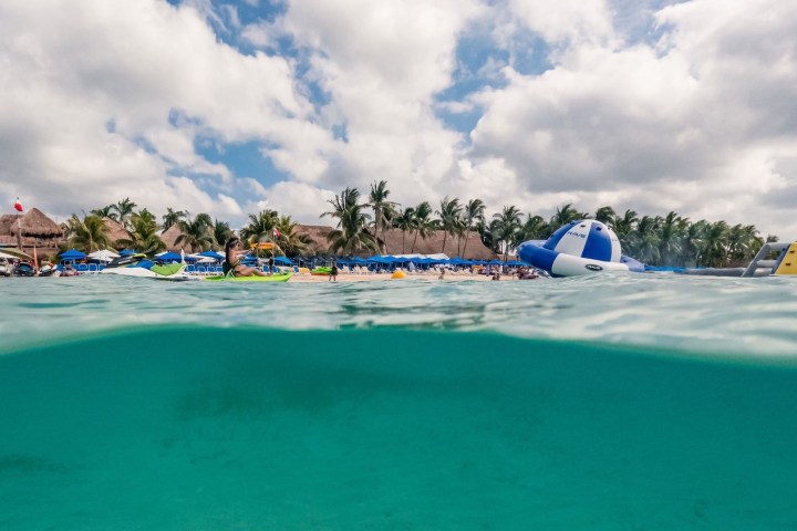 Beach scene with clear blue water, palm trees, and inflatable water toys under a cloudy sky.