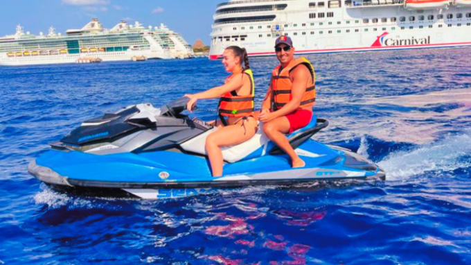 Two people on a blue jet ski in the ocean with cruise ships in the background.