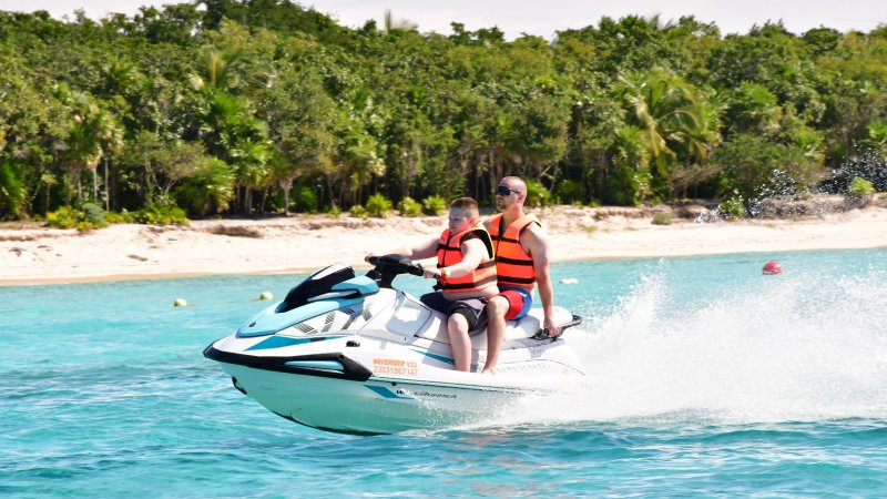 Two people in life vests riding a jet ski on clear blue water near a sandy beach with trees.