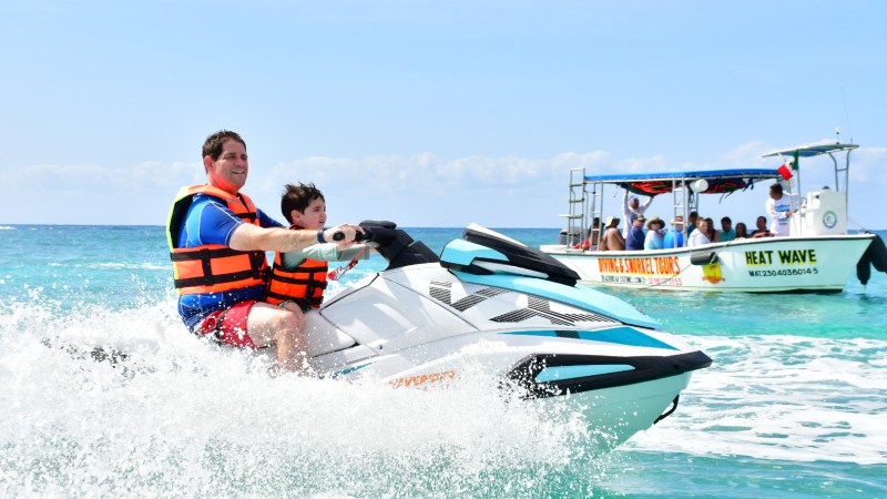 Two people riding a jet ski on the ocean with a tour boat in the background.